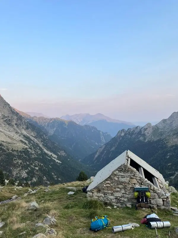 Stone mountain hut with sleeping mats outside and a valley behind